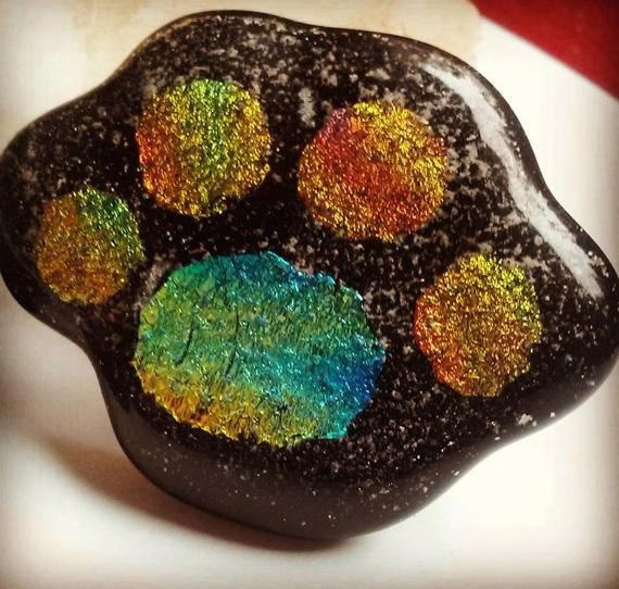 Handcrafted paw print cremation stone made with cremation ash, featuring a glossy black memorial glass base with iridescent multicolor paw pads and delicate ash inclusions throughout.