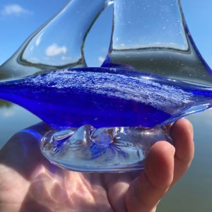 A close-up of a clear glass sailboat sculpture, showing the cobalt-blue glass base infused with cremation ash held between the artist’s fingers. Sunlight highlights the texture of the ash within the blue glass and the smooth contours of the clear glass sails.