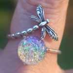 Dragonfly Cremation Ring in clear rainbow color variation featuring a sparkling iridescent memorial glass stone set beside a detailed silver dragonfly, shown in a close-up outdoor view