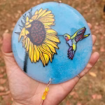 Handheld round glass suncatcher with a painted sunflower and hummingbird on a blue background, infused with cremation ash and finished with dangling crystal accents.