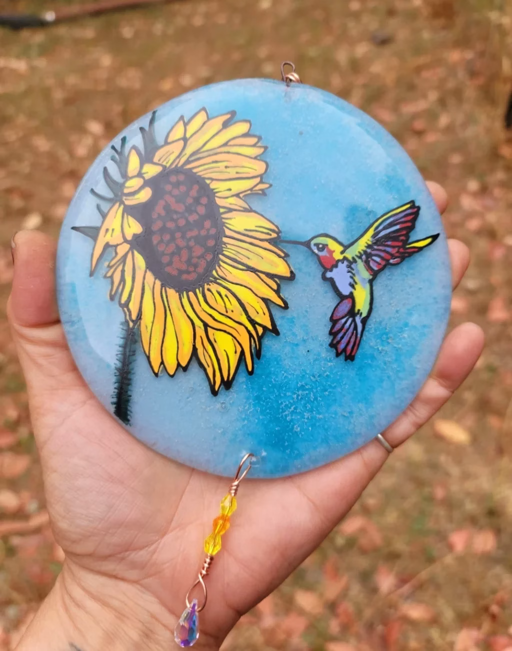 Handheld round glass suncatcher with a painted sunflower and hummingbird on a blue background, infused with cremation ash and finished with dangling crystal accents.