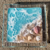 Oceanside Ring Dish with Cremation Ash showing a square clear glass dish with turquoise ocean water, white foaming wave detail, sandy shoreline, and embedded seashells viewed in natural sunlight
