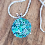 Sterling silver halo cremation pendant with aqua crinkle glass resting on a wooden surface.