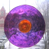 Circular memorial glass suncatcher containing cremation ash, featuring rippled purple glass with a glowing amber center, hanging in a window with soft daylight and an outdoor winter scene visible in the background.