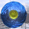 Circular memorial glass suncatcher containing cremation ash, featuring rippled deep blue glass with a translucent yellow center, hanging in a window and illuminated by natural light with a winter landscape visible in the background.