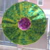 Round memorial glass suncatcher containing cremation ash, featuring vibrant rippled green glass with a rich purple center, suspended in a window with a softly blurred urban building visible in the background.