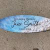Oceanside Memorial Surfboard with cremation ash, featuring ocean blue wave texture, white shoreline details, embedded seashells, and personalized memorial text, shown horizontally on a concrete surface