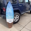 Oceanside Memorial Surfboard made with cremation ash, featuring layered ocean blue and white wave design over natural wood tones, shown leaning against a vehicle outdoors
