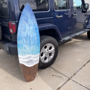 Oceanside Memorial Surfboard made with cremation ash, featuring layered ocean blue and white wave design over natural wood tones, shown leaning against a vehicle outdoors