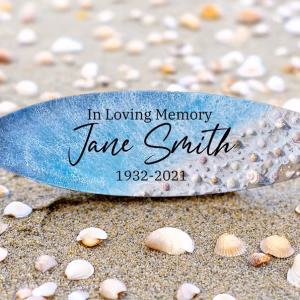 Memorial surfboard with cremation ash display reading In Loving Memory Jane Smith 1932–2021 resting on sand surrounded by seashells