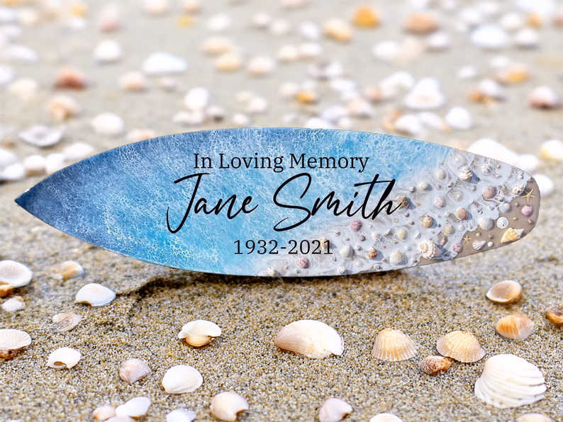 Memorial surfboard with cremation ash display reading In Loving Memory Jane Smith 1932–2021 resting on sand surrounded by seashells