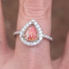 Front-facing close-up of a teardrop-shaped cremation ring worn on a finger. The red-orange memorial stone, infused with ashes and showing shimmering color shifts, is set in a pear-shaped prong setting. It is surrounded by a halo of clear cubic zirconia stones, with additional CZ stones lining the slim band.
