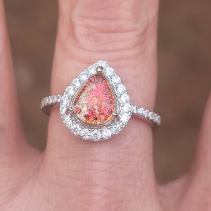 Front-facing close-up of a teardrop-shaped cremation ring worn on a finger. The red-orange memorial stone, infused with ashes and showing shimmering color shifts, is set in a pear-shaped prong setting. It is surrounded by a halo of clear cubic zirconia stones, with additional CZ stones lining the slim band.