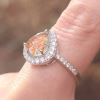 A close-up photo of a teardrop-shaped cremation ring in red-orange, shown on a finger. The pear-shaped memorial glass stone—infused with ashes—displays shimmering red, orange, and gold tones. It is framed by a halo of clear gemstones, with additional stones set along the slim band. The image highlights the ring’s sparkle, prong setting, and raised profile in soft natural light.
