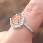 A close-up photo of a teardrop-shaped cremation ring in red-orange, shown on a finger. The pear-shaped memorial glass stone—infused with ashes—displays shimmering red, orange, and gold tones. It is framed by a halo of clear gemstones, with additional stones set along the slim band. The image highlights the ring’s sparkle, prong setting, and raised profile in soft natural light.