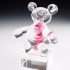 A clear glass teddy bear sculpture displayed on a transparent square base, featuring a pink swirling core that contains incorporated cremation ash.