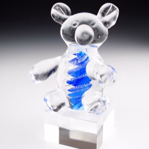 A clear glass teddy bear sculpture sitting atop a transparent square base, featuring a vibrant blue swirling core that contains cremation ash.