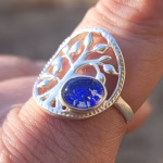 Close-up of a Family Tree cremation ring on a finger, featuring a silver tree-of-life design with a round sparkling blue memorial glass stone containing cremation ash.