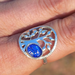 Close-up of a Family Tree cremation ring worn on a finger, featuring a silver tree design with a sparkling blue memorial glass stone containing cremation ash.