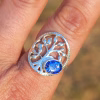 Front view of a Family Tree cremation ring in sterling silver worn on a finger, featuring an oval sparkling blue memorial glass stone containing cremation ash set beside an open tree design.