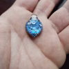 Handcrafted blue dichroic glass cremation pendant resting in the palm, featuring shimmering metallic blue tones and scattered white flecks of encased cremation ashes beneath a clear glass cap, shown close-up to highlight texture and size