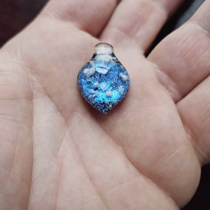 Handcrafted blue dichroic glass cremation pendant resting in the palm, featuring shimmering metallic blue tones and scattered white flecks of encased cremation ashes beneath a clear glass cap, shown close-up to highlight texture and size