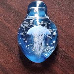 Handcrafted glass jellyfish cremation pendant featuring a detailed white jellyfish suspended in vibrant blue glass with swirling white flecks of encased cremation ashes, shown in a full top-down view to highlight shape, color, and clarity