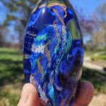 Close-up of the Reflections Glass Paperweight with cremation ash, showing deep cobalt and teal glass with flowing silver ash details and a twisted blue-and-gold ribbon design, held outdoors in natural light.