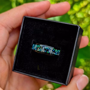 Close-up of a blue opal cremation ring made with ashes, displayed inside a black ring box held outdoors against vibrant green foliage.