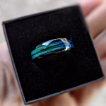 Hand holding a black ring box containing a blue opal memorial ring made with hair, featuring vibrant teal and sapphire inlay bands.