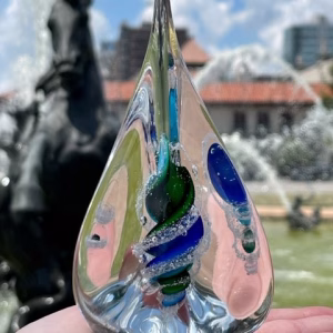 Pyramid Memorial with Cremation Ash in aqua, blue, and green glass held in hand with city fountain background
