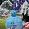 Round aqua glass memorial vase with cremation ash holding purple flowers, photographed outdoors near a fountain.