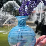 Round aqua glass memorial vase with cremation ash holding purple flowers, photographed outdoors near a fountain.