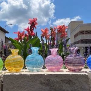 Six round glass memorial vases in assorted colors displayed outdoors on a stone ledge.