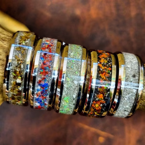 Cremation rings with colored stone inlay arranged on a natural antler, showcasing multiple polished metal bands filled with ash-infused stone inlays in neutral, red, blue, green, opalescent, and multicolor variations.