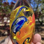 Handheld oval glass paperweight with flowing amber, blue, and yellow patterns surrounding cremation ash, shown outdoors in natural light.