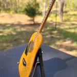 Side view of a glass memorial guitar incorporating cremation ash, displayed on a small stand outdoors with a softly blurred natural background.