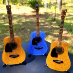 Three glass memorial guitars incorporating cremation ash displayed outdoors, shown in blue and gold color variations on individual stands.
