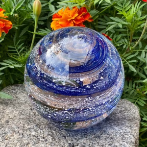 Hand-blown Infinite Staircase cremation paperweight featuring swirling bands of Sparkle Blue and Sienna Brown glass with cremation ashes suspended throughout. Displayed outdoors on a stone surface with vibrant marigold flowers and greenery in the background.