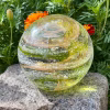Hand-blown Infinite Staircase cremation paperweight featuring swirling layers of yellow gold sparkle mixed with granny apple green, infused with cremation ashes throughout. The glass sphere rests on an outdoor stone surface with marigolds and green foliage in the background.