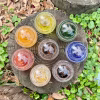 Top-down view of eight Infinite Staircase cremation paperweights arranged in a circle on a tree stump, each in a different vibrant color. The glass spheres display swirling designs with cremation ashes suspended inside, surrounded by green groundcover and fallen leaves.