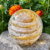 Hand-blown glass Infinite Staircase cremation paperweight in gold sparkle, featuring swirling golden ribbons with visible cremation ashes suspended inside, displayed outdoors on a stone surface with marigold flowers and green foliage in the background.