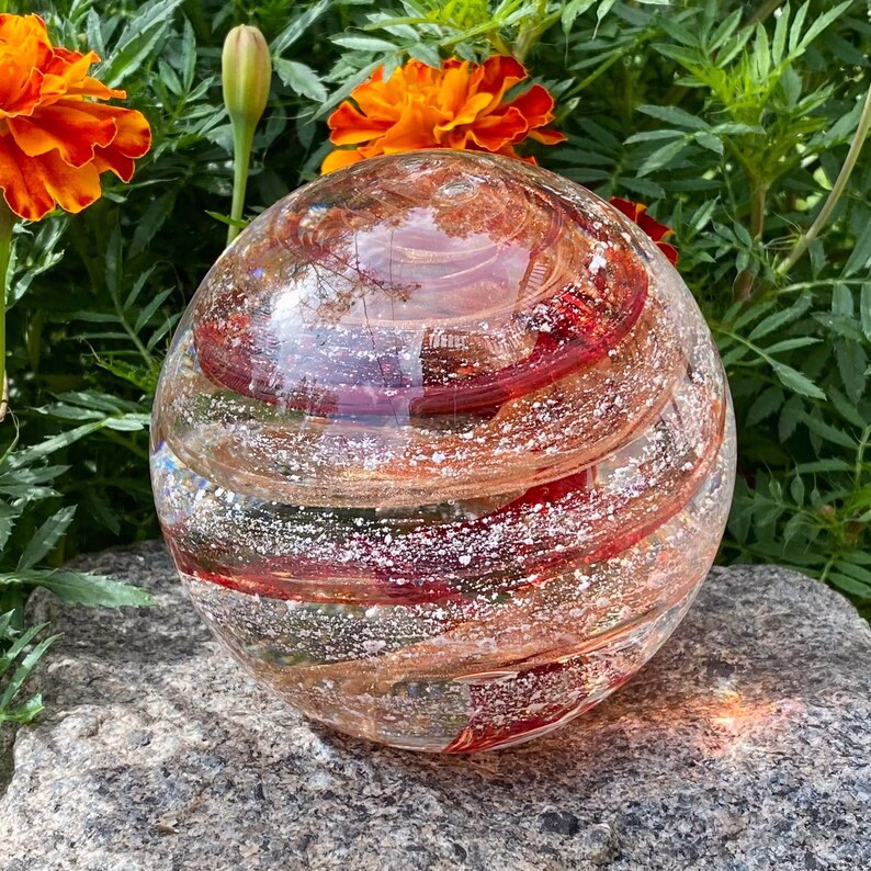 Hand-blown Infinite Staircase cremation paperweight featuring swirling bands of Copper Ruby and Sparkle Gold glass with cremation ashes suspended throughout. Displayed outdoors on a stone surface with vibrant marigold flowers and green foliage in the background.