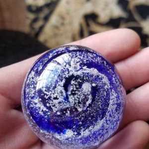 Close-up of a blue memorial prayer stone made with cremation ash, showing delicate ash inclusions suspended in layered glass and a smooth, comforting shape resting in the palm of a hand.