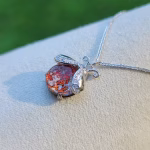 A red orange opal Ladybug Cremation Pendant displayed on a soft beige jewelry stand, shown from a side angle to highlight the shimmering memorial stone and silver wing details.