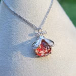 Red ladybug cremation pendant featuring shimmering red dichroic memorial glass with ashes, set in a silver ladybug design and displayed on a necklace stand outdoors.