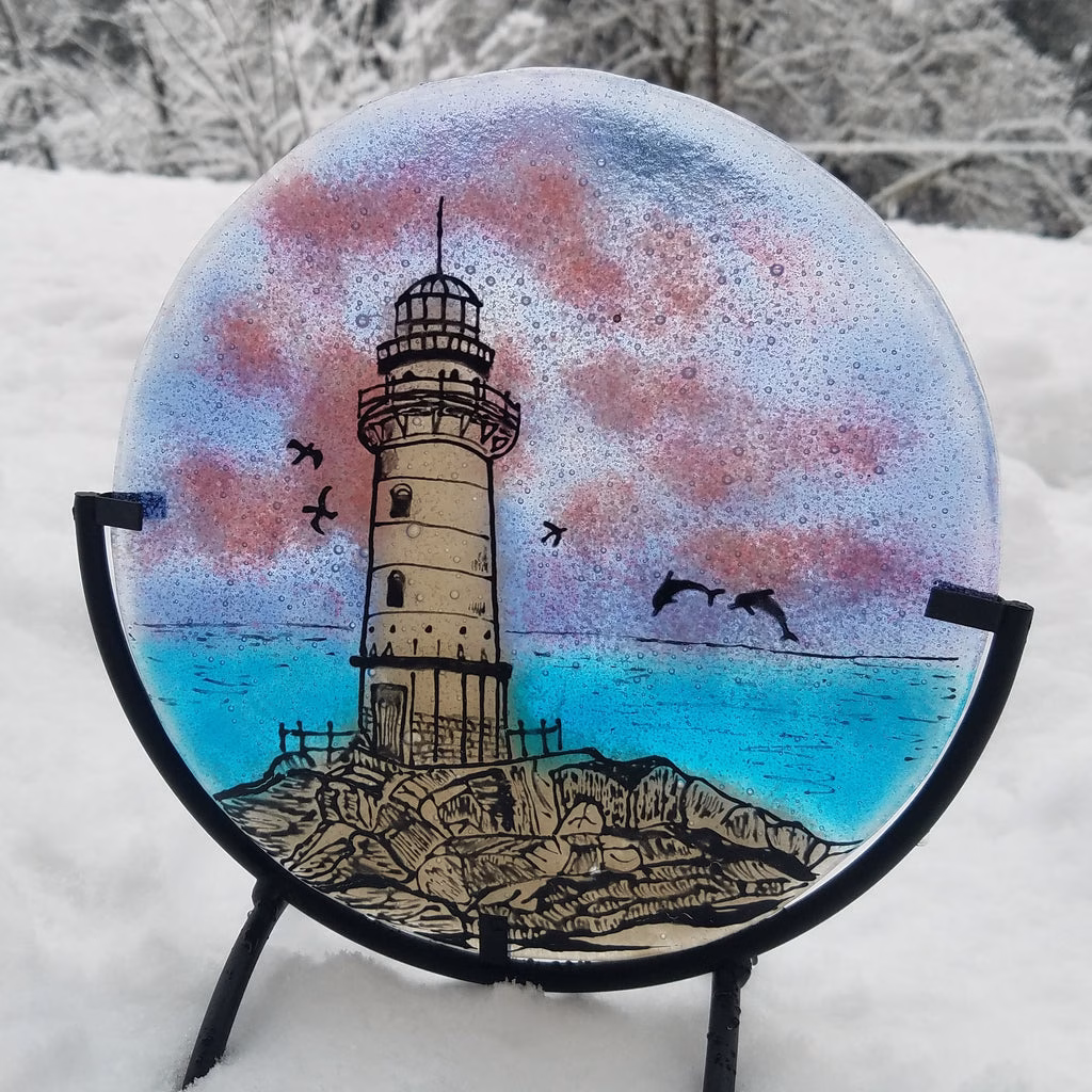 Round glass lighthouse suncatcher infused with cremation ash, featuring a hand-painted coastal scene with rocky shoreline, blue ocean, pink clouds, flying birds, and dolphins, displayed outdoors in fresh snow.