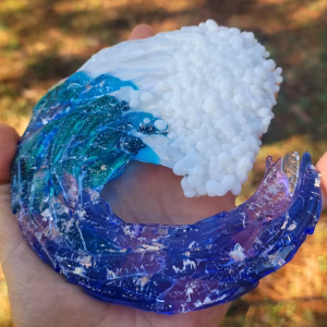 Ocean Wave Suncatcher with Cremation Ash held in hand, showcasing layered blue and teal glass wave with deep curl, textured white sea foam crest, and memorial ash fused throughout the handcrafted glass