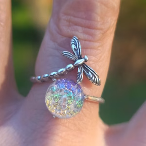 Dragonfly Cremation Ring in the clear rainbow color variation, shown in natural sunlight with a sparkling iridescent memorial glass center and a detailed silver dragonfly wrapped around the band