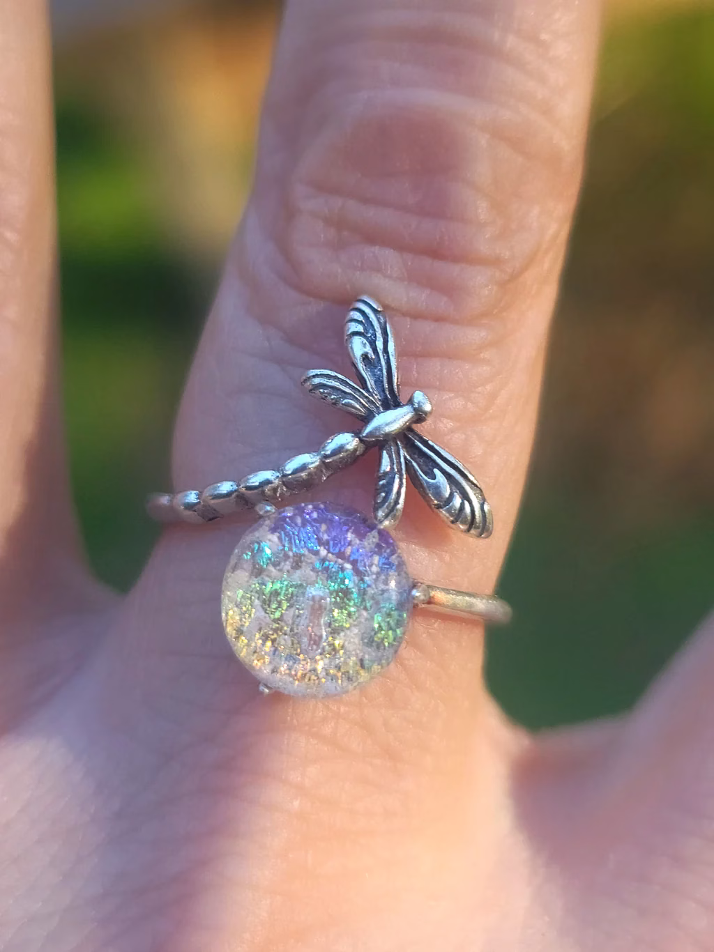 Dragonfly Cremation Ring in the clear rainbow color variation, shown in natural sunlight with a sparkling iridescent memorial glass center and a detailed silver dragonfly wrapped around the band